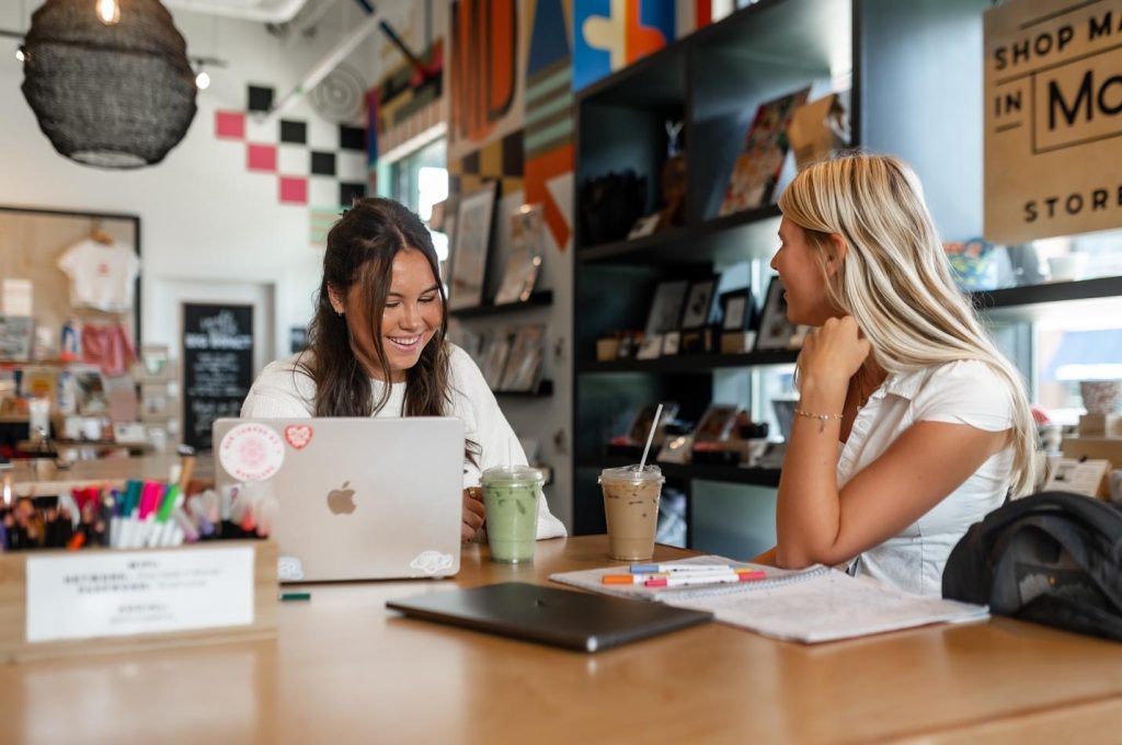 Two women sit at a table in a colorful cafe, talking and smiling as they enjoy making friends at UMD. One has a laptop and a green smoothie, while the other has iced coffee and notebooks. The background shows shelves with books and decor.