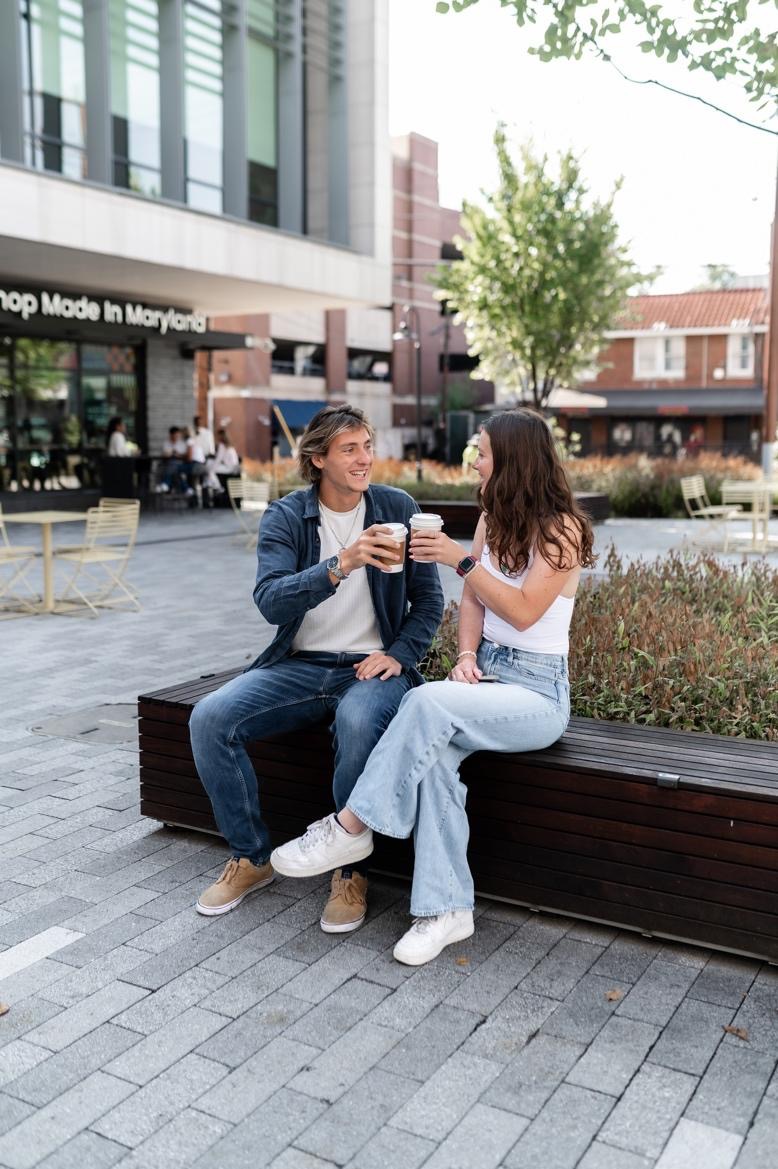 A man and woman sit on a bench outdoors, smiling and holding coffee cups. They appear to be making friends at UMD, enjoying a casual conversation in an urban plaza with shops and trees in the background.