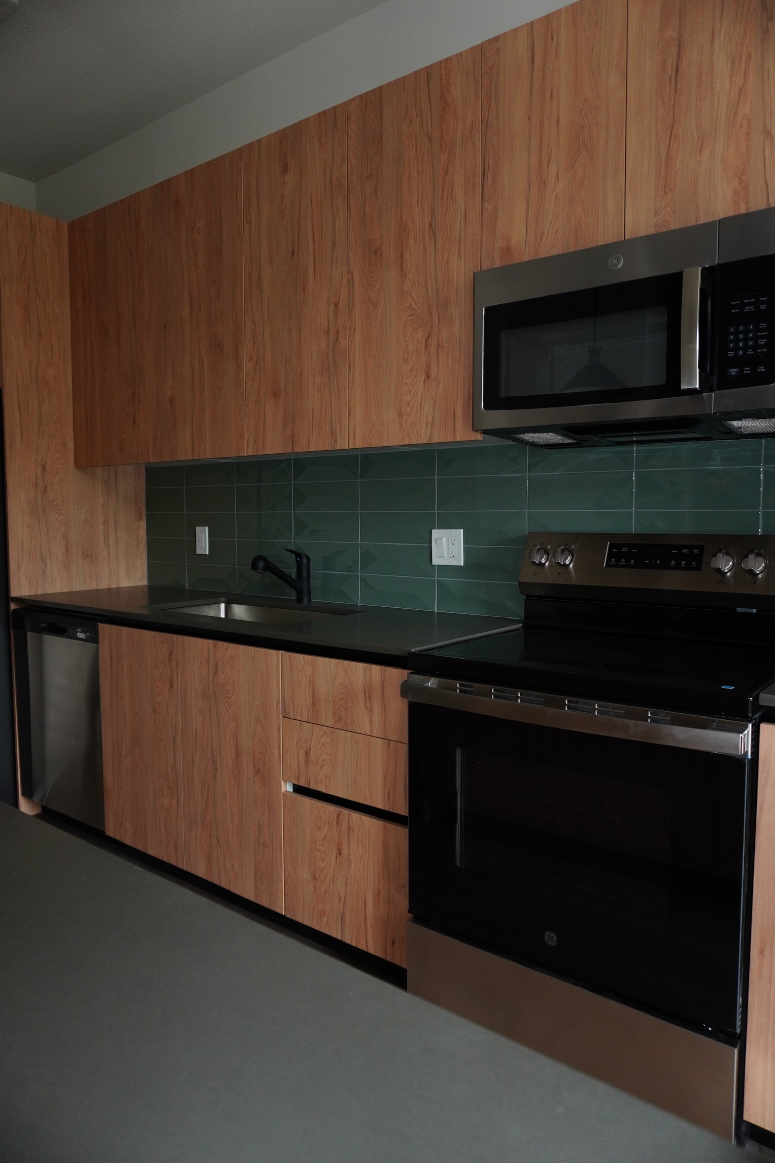 Modern kitchen with wood cabinets, green tile backsplash, black countertop, stainless steel oven, microwave, dishwasher, and a black sink faucet. Lighting is dim, giving the room a cozy and sleek appearance.