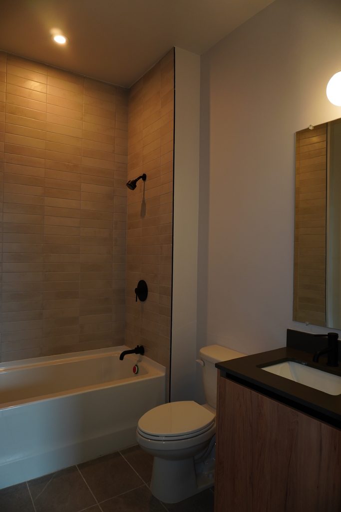 A modern bathroom with beige tile walls, a bathtub with a black shower fixture, a toilet, and a vanity with a black countertop and sink under a wall-mounted mirror, softly lit by ceiling and mirror lights.