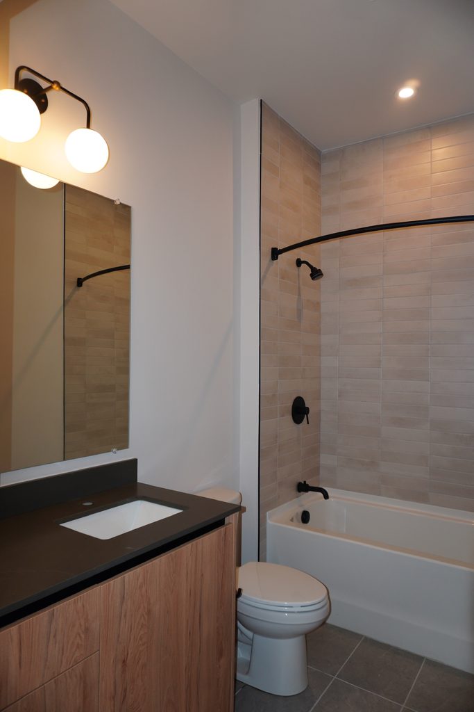 Modern bathroom with wood vanity, sink, and black countertop, a toilet, and a bathtub with beige tile surround. Black fixtures and lighting, plus a large mirror, complete the clean, minimalist design.