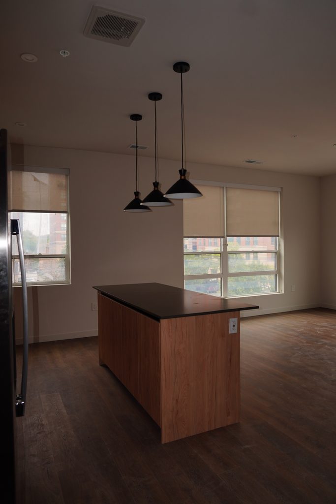 A modern kitchen with a wooden island, three black pendant lights above, large windows with beige blinds, and dark wood flooring.