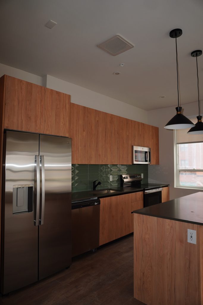 Modern kitchen with wood cabinets, stainless steel appliances, green tile backsplash, black pendant lights, and a large island with a dark countertop. Natural light comes in from a large window.