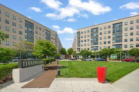 A grassy courtyard is surrounded by two tall apartment buildings with many windows, trees, parked cars, a red sculpture, and a sign that reads TERRA.