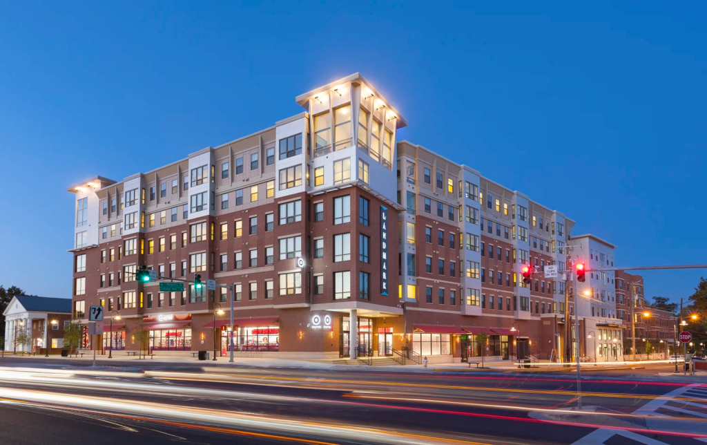 Modern five-story apartment building with retail stores on the ground floor, including a Target, photographed at dusk with car light trails and illuminated windows. Traffic lights and street signs are visible in front.