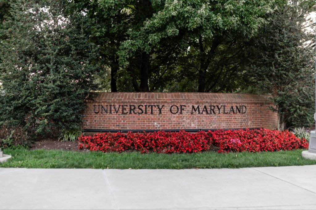 A brick sign reads University of Maryland, surrounded by red flowers and green trees, with a sidewalk in front.