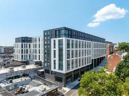 Modern multi-story apartment building with large windows and geometric white and dark gray facade, set against a clear blue sky with a single cloud and some trees in the foreground.