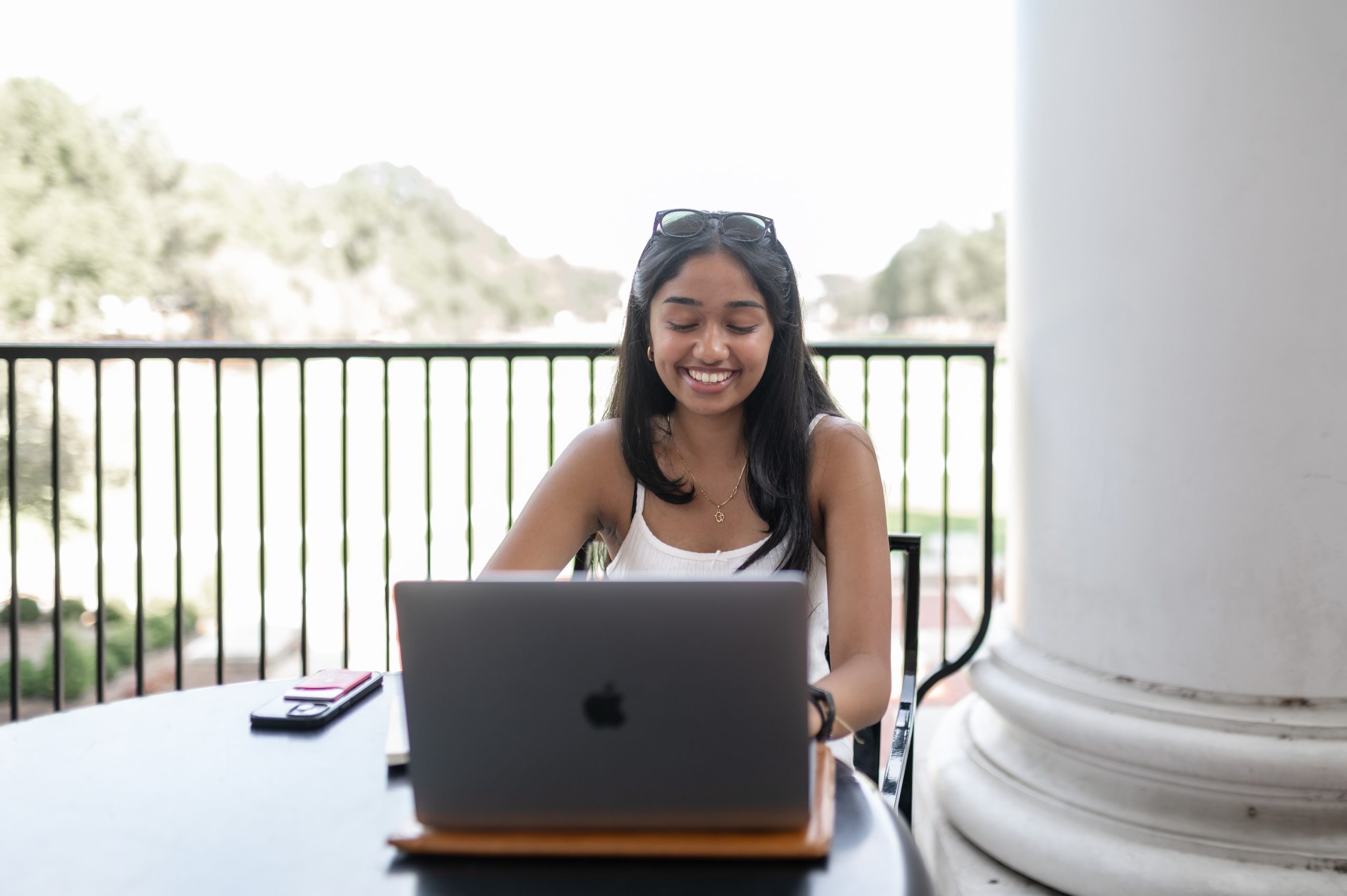 A smiling woman sits at a table outdoors using a laptop.
