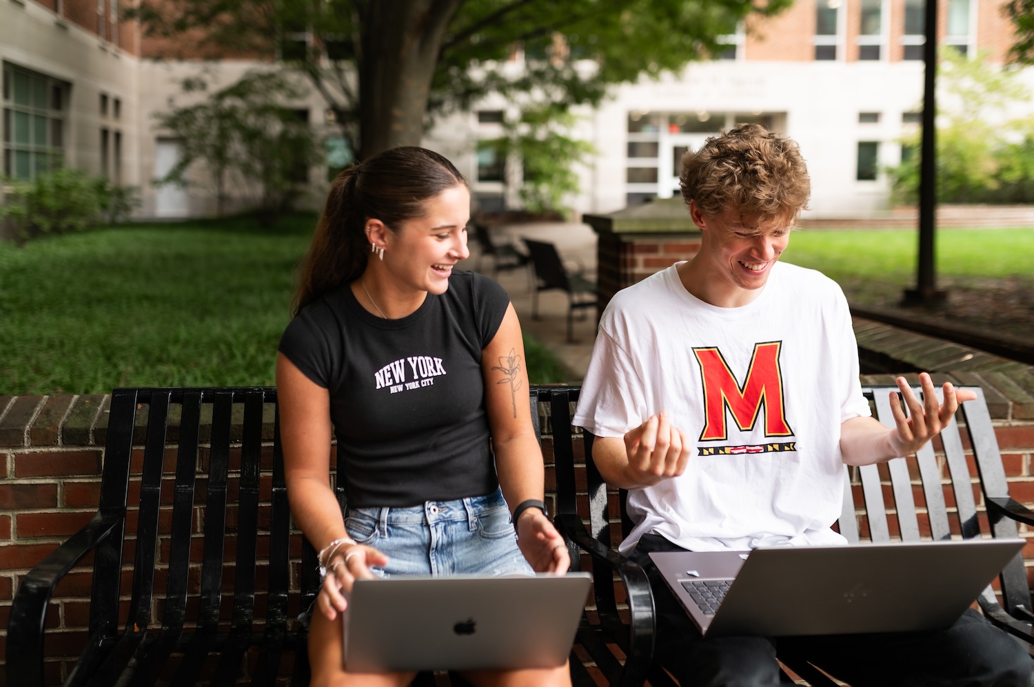A young man and woman laughing while working on laptops together on a park bench.